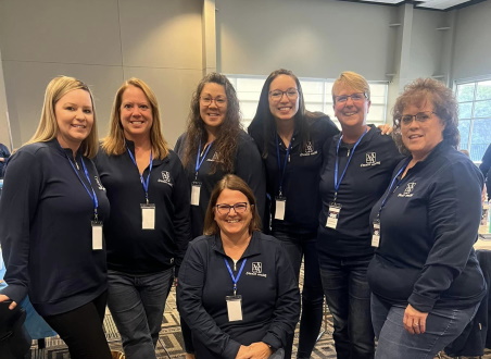 7 females standing together wearing navy blue sweaters and jeans